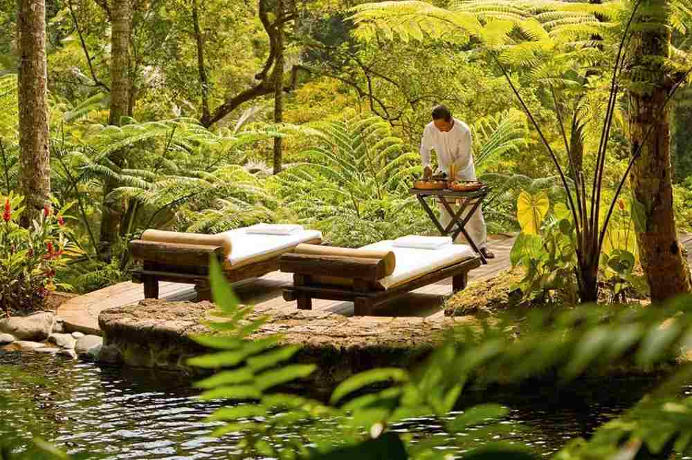 Outdoor wellness spa massage setup in lush tropical rainforest with bamboo loungers and therapist preparing treatments